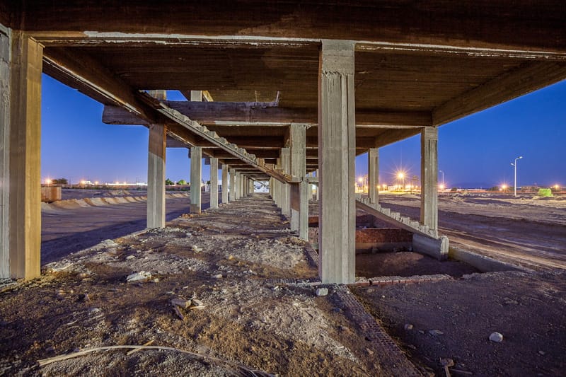 Beneath the deck of an unfinished building at dusk, pillars catching warm light, city lights in the distance
