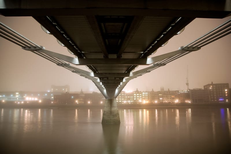 Beneath the Millennium Bridge in fog, city lights strung along the far bank