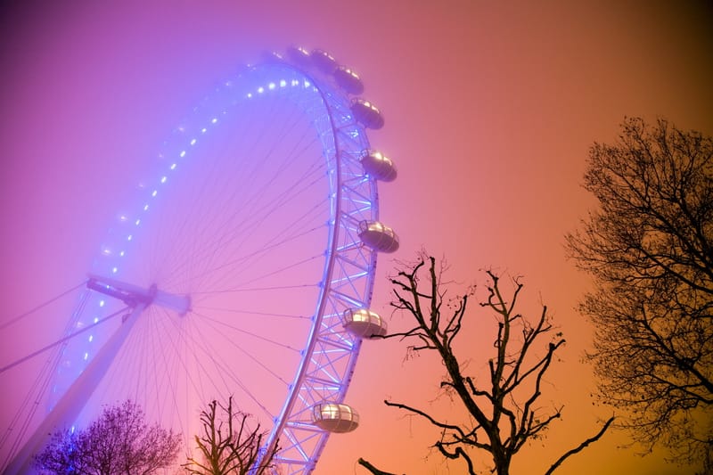 The London Eye lit blue-violet in fog behind bare trees, orange night sky