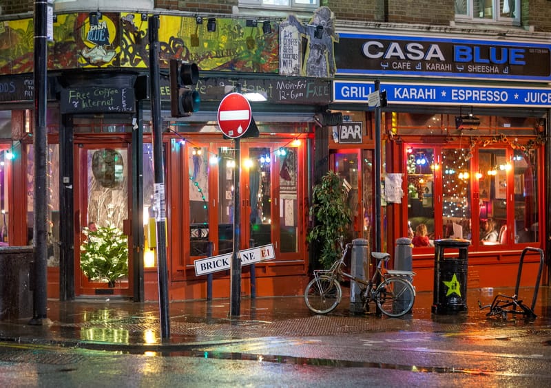 Rain-soaked Brick Lane at night — red café front, wet pavement reflections, bicycles at the kerb