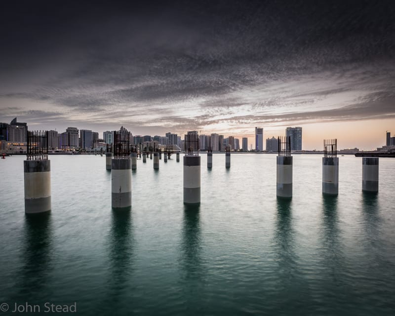 Twin rows of weathered pier columns standing in the sea, a high-rise skyline behind, UAE