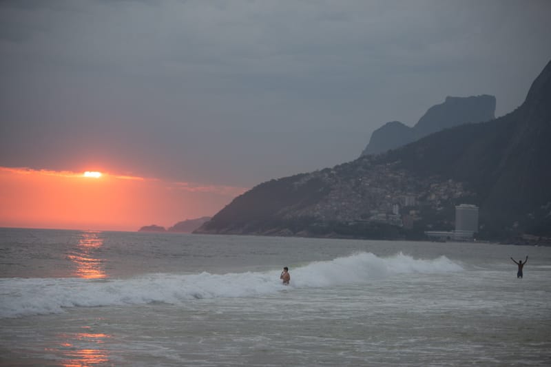 A red sun setting into the sea off Ipanema, a swimmer raising their arms in the surf