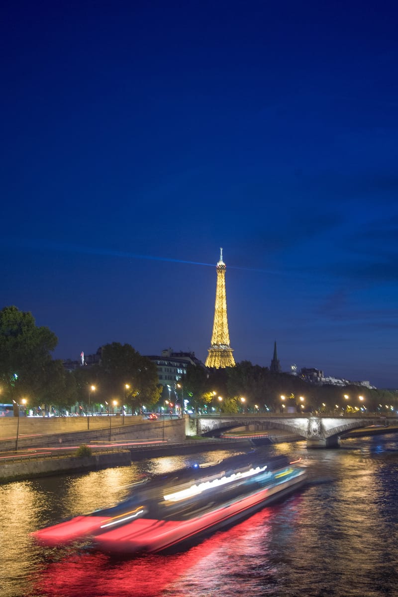 A boat's light trail sweeping down the Seine at night, the Eiffel Tower golden beyond