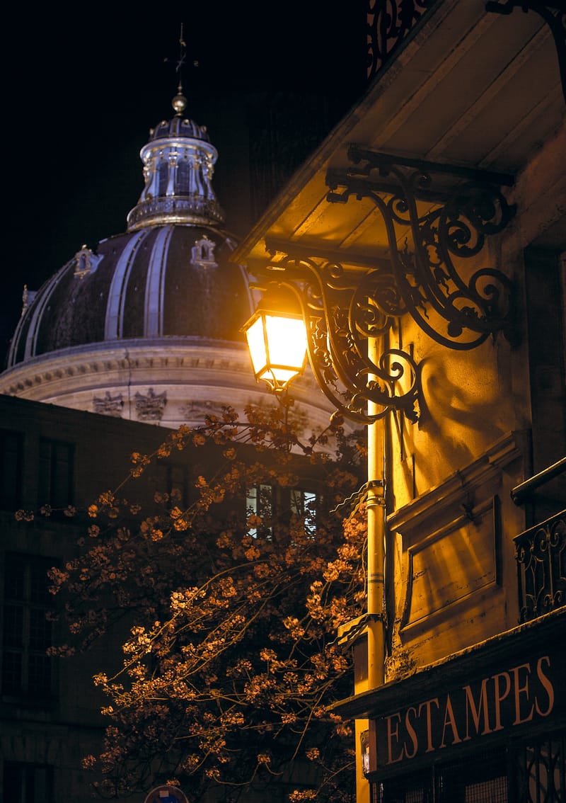 A lantern glowing beside an old print-shop front, the dome of the Institut de France behind, Paris