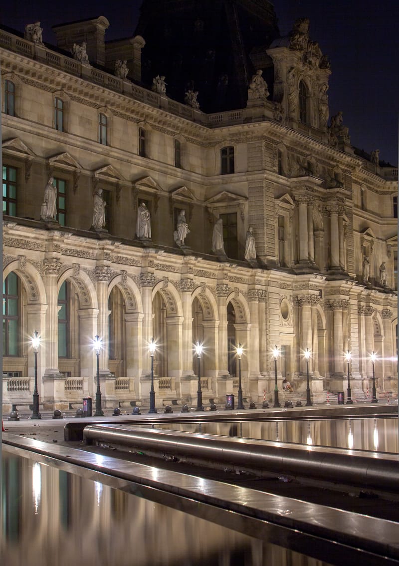 The floodlit façade of the Louvre at night, fountain pools in the foreground