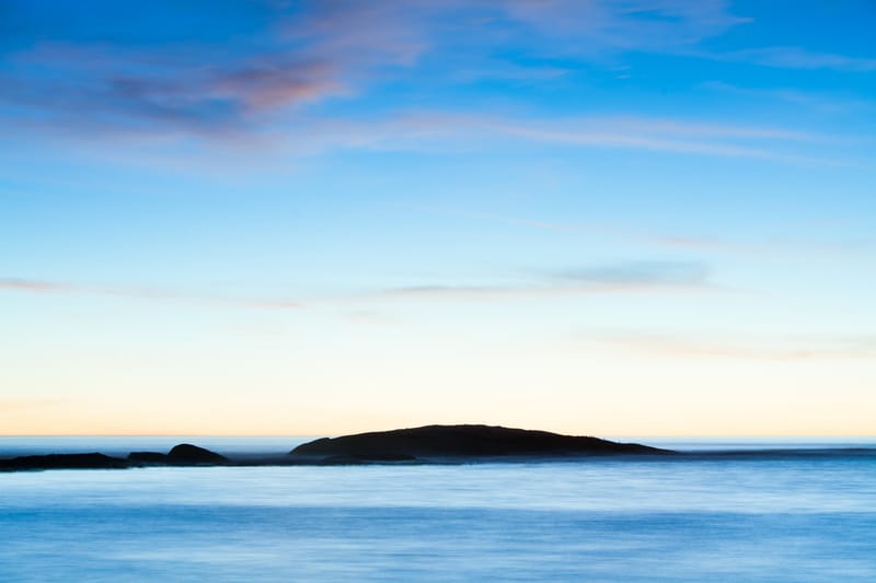 A small island silhouetted offshore under drifting clouds, silky long-exposure sea
